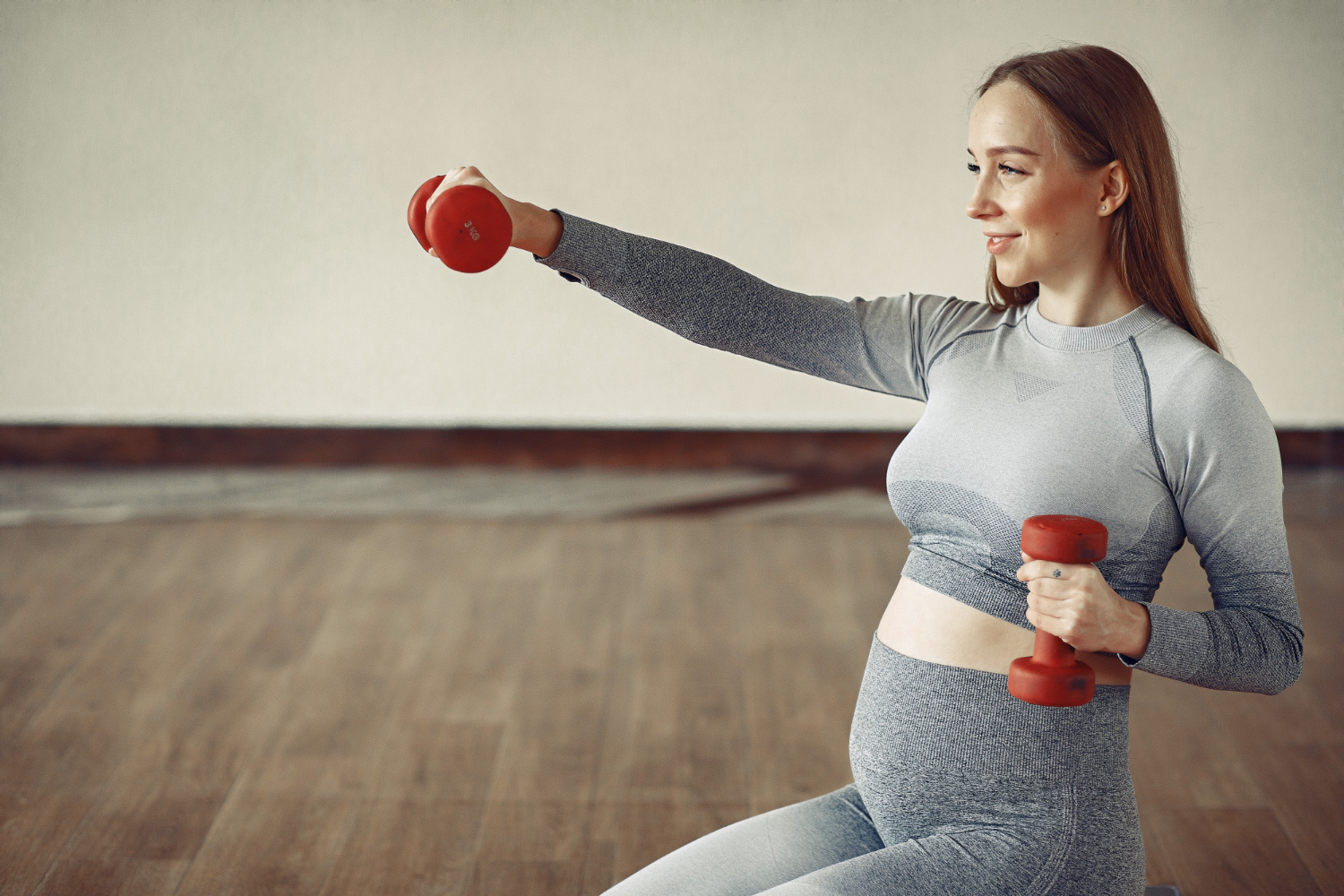 A pregnant woman in grey athletic wear lifting red dumbbells in a gym with wooden floors.